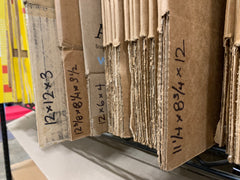 Re-used cardboard boxes labeled with their three dimension sizes, folded flat, and stacked on a shelf for easy categorization