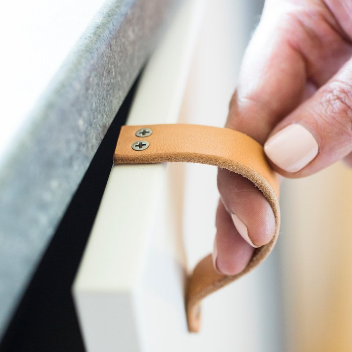 Woman is pulling open a bottom cabinet door under sink with a natural leather Lovejoy handle. On top of the cabinet is the two tiny wood screws that attach the handle to the IKEA cabinet door.