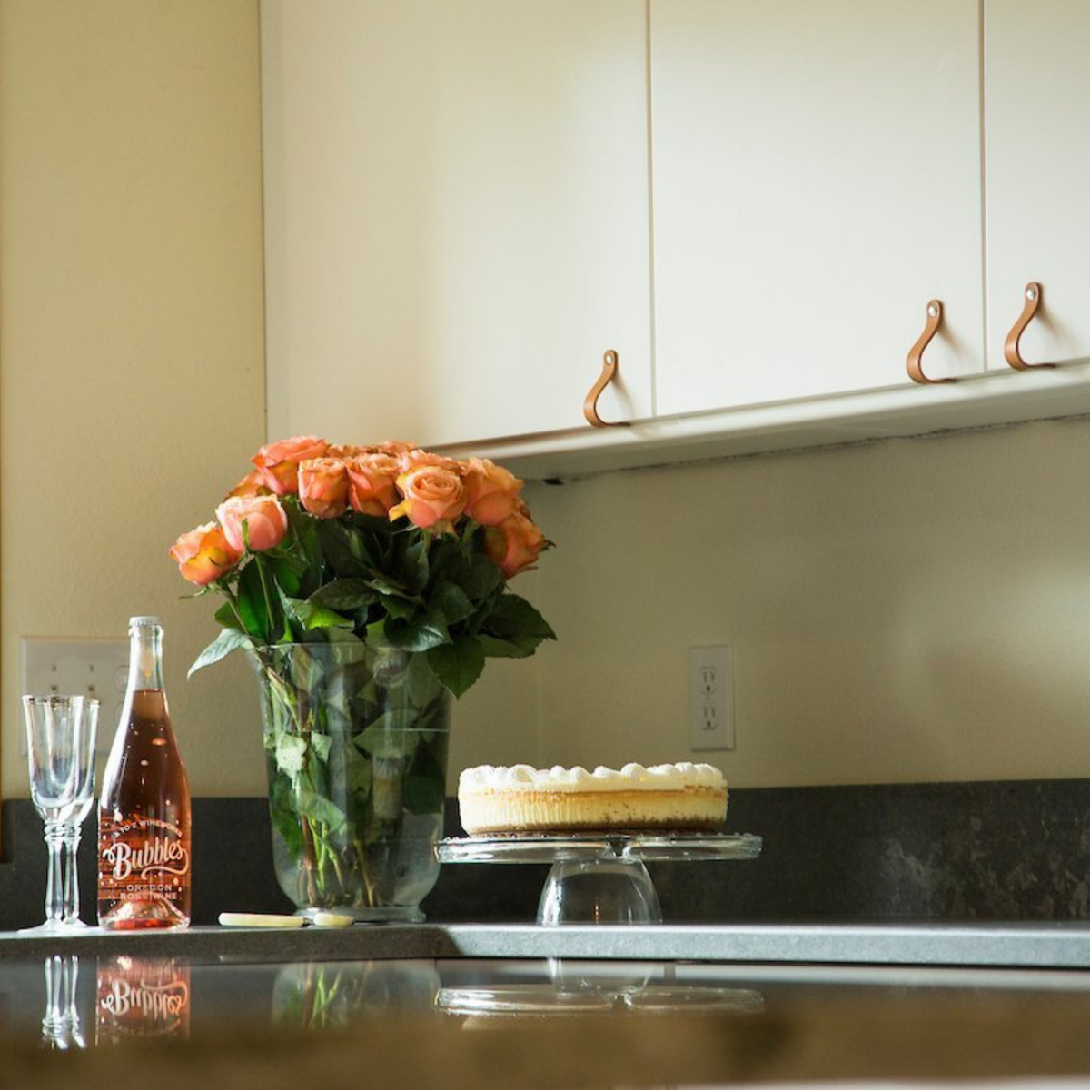 White kitchen with white walls, white cabinets, and gray concrete countertop with soothing tan natural vegetable-tanned leather Lovejoy handles on the cabinet doors.  With a birthday cake, pink roses, and rose champagne. 