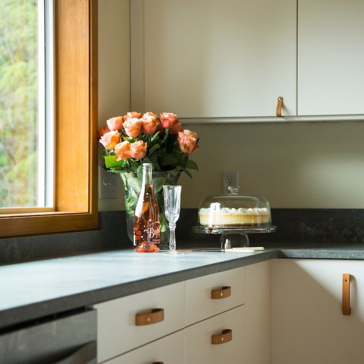 White kitchen with a beautiful bouquet of pink roses, rose champagne, and cake with light gray countertops and natural vegetable-tanned leather handles