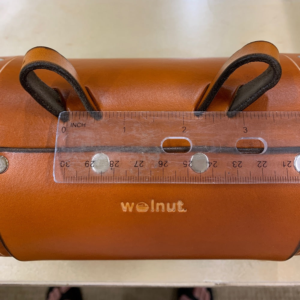 Honey leather variant of bicycle barrel bag being measured between two connecting points with clear ruler. The ruler shows that the difference between the two leather loops is three inches.