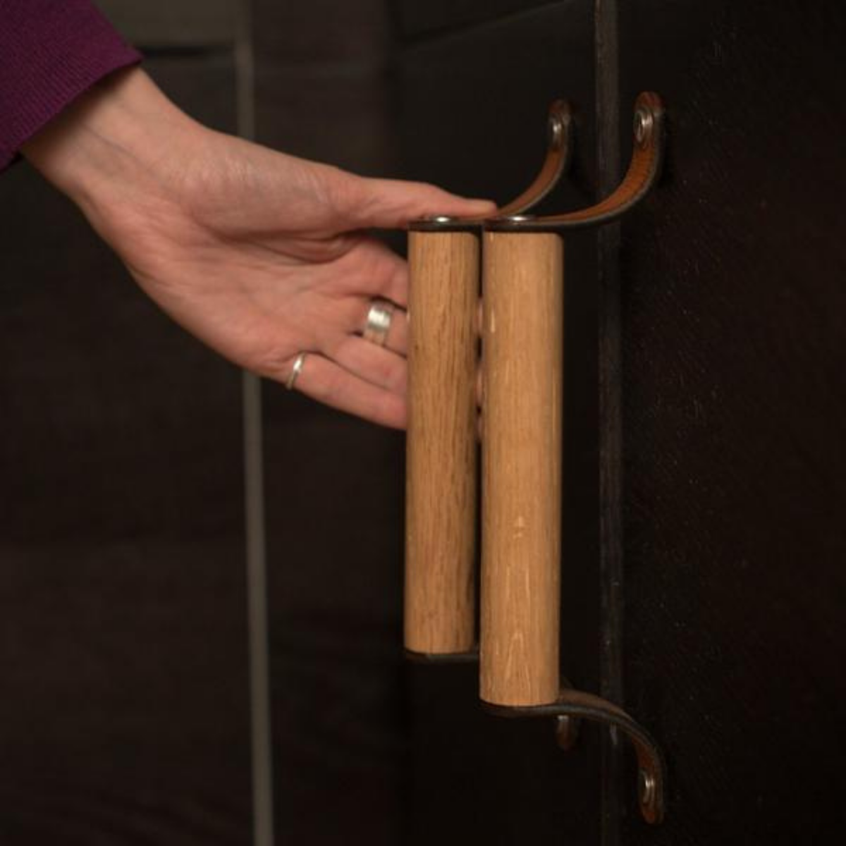 Woman reaching down to a white oak wood and honey leather handle on black-brown dark kitchen cabinetry in an IKEA kitchen.