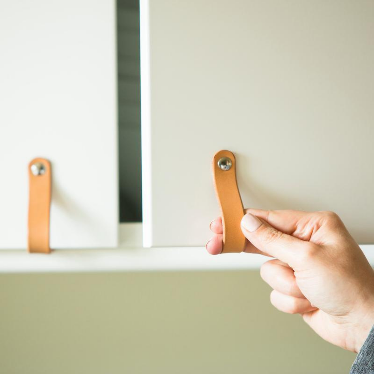 A woman&#39;s hand is reaching towards a tan, natural leather cabinet handle on a set of white cabinets. The handle loops around the bottom of the European style (IKEA) cabinet door and attaches on the underside.