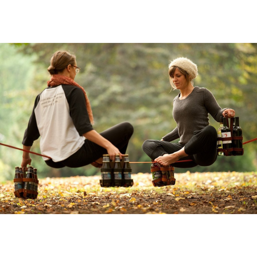 Two women balancing on a slackline in a park in Portland Oregon brewery town, holding leather 6-packs of beer as balance weights