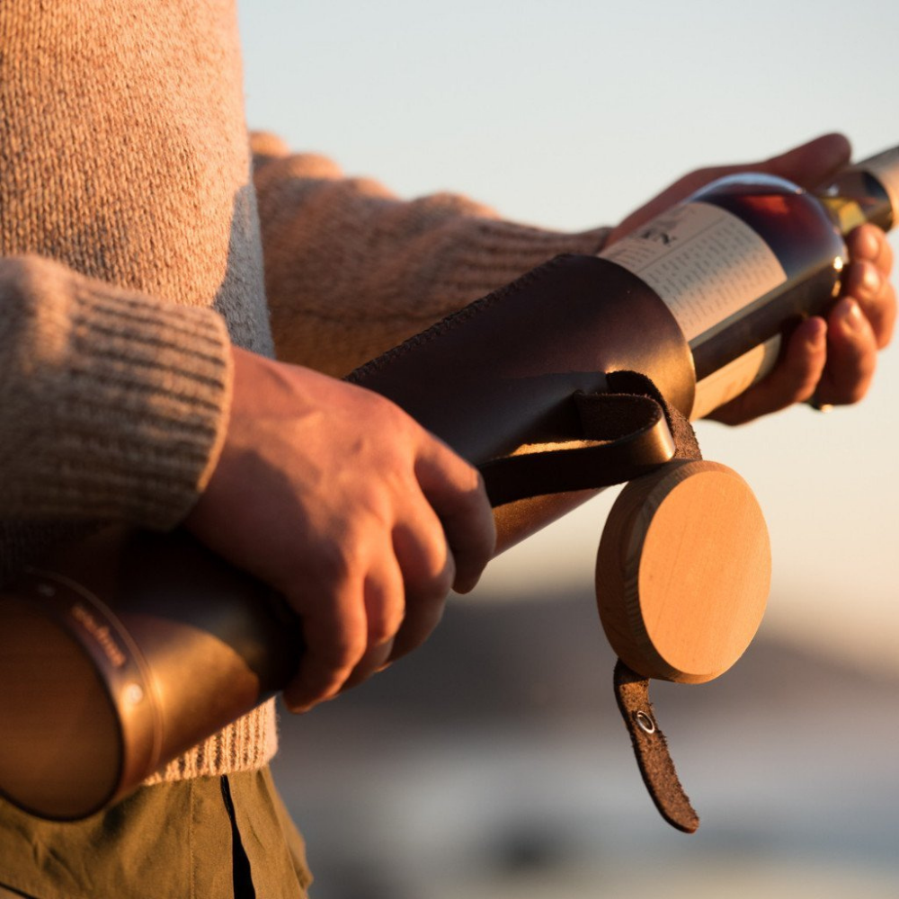 A man pulling a bottle of Scotch whiskey out of a hand-stitched leather case at sunset on the beach. The case&#39;s wood lid is flapped open but still attached to the case with a leather strap. 