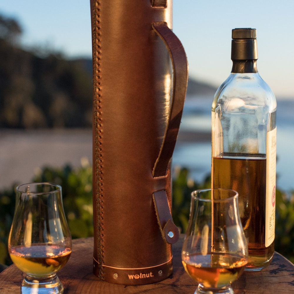 Still life scene of a leather whiskey bottle case, a bottle of Scotch, and two filled Glencairn glasses on a beautiful Oregon Coast beach with the blue sky in the background. 