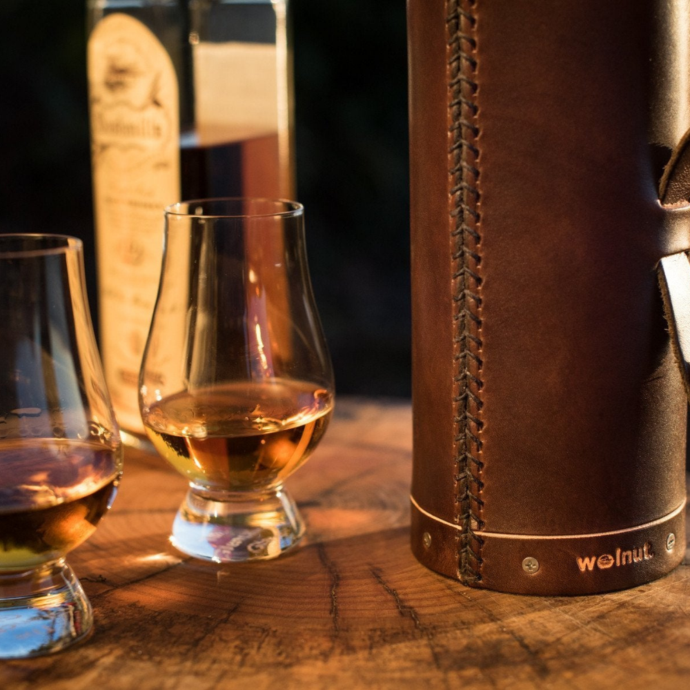 Close-up detail of the leather whiskey bottle case showing the bottom half of the case. Around the bottom of the case is a grooved out line showing the natural color of the leather below the dark brown hand-dyed surface and the &quot;Walnut&quot; logo.