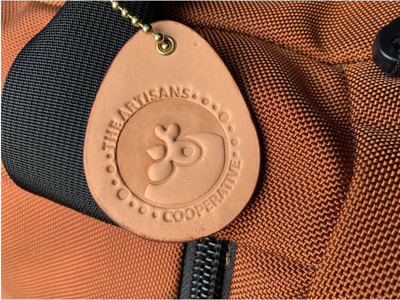 Close-up of the egg-shaped leather fundraiser keychain for Artisans Cooperative attached to a orange luggage bag with brass ball chain as a luggage identifier tag