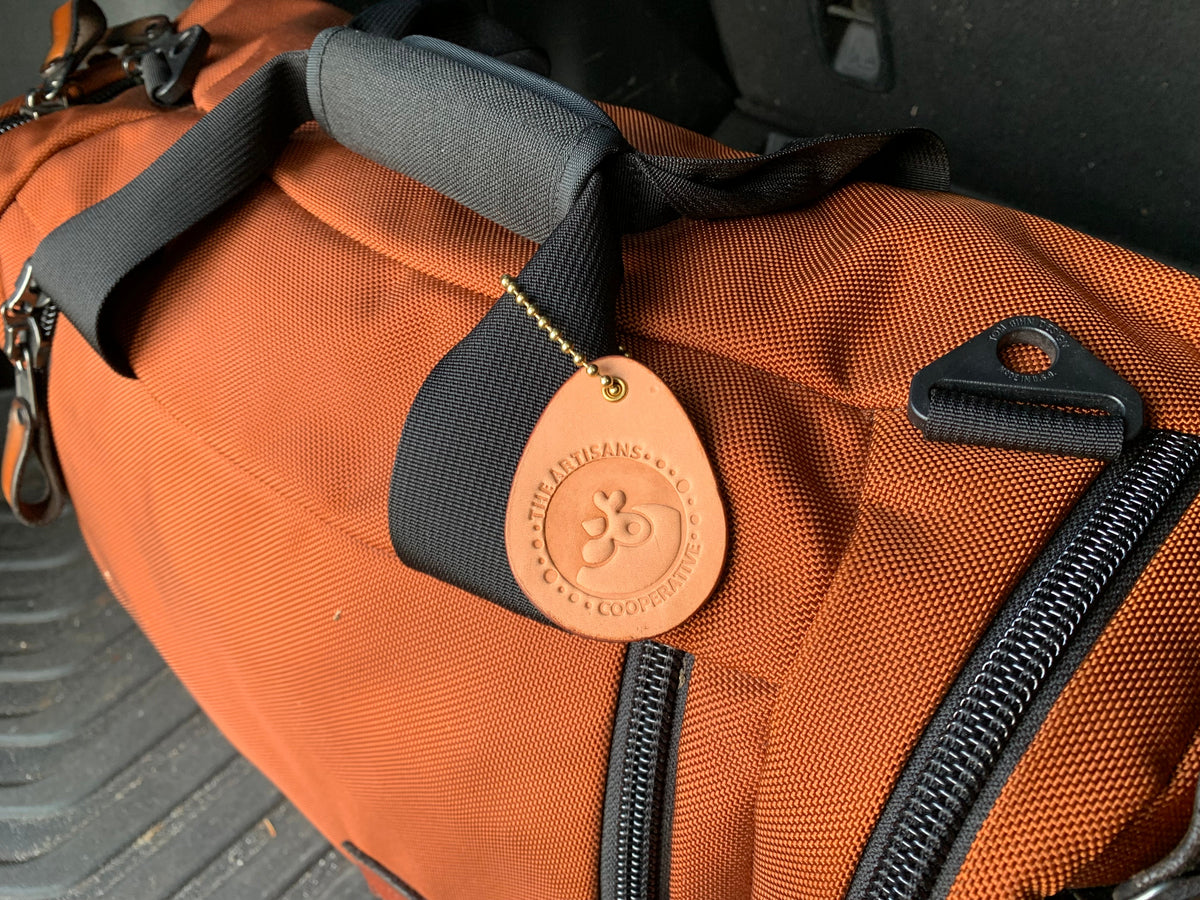 Close-up of the egg-shaped leather fundraiser keychain for Artisans Cooperative attached to a orange luggage bag with brass ball chain as a luggage identifier tag