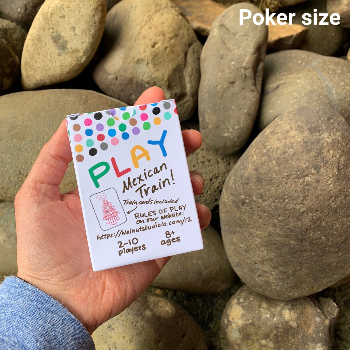 A hand holding a set of double 12 domino playing cards . The cards take up the entire woman&#39;s hand. The backside of the box says &quot;Play Mexican Train!&quot; The cards are standard playing card size. 