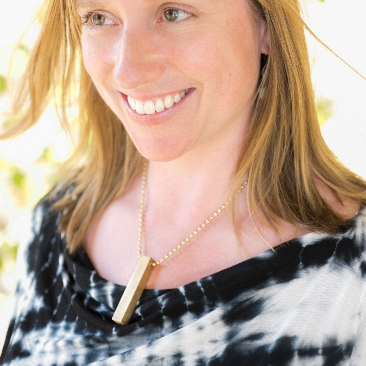 Close-up of a woman&#39;s neck and collarbone, wearing the square brass travel dice as a necklace using a longer section of brass beaded chain.  The woman is smiling.